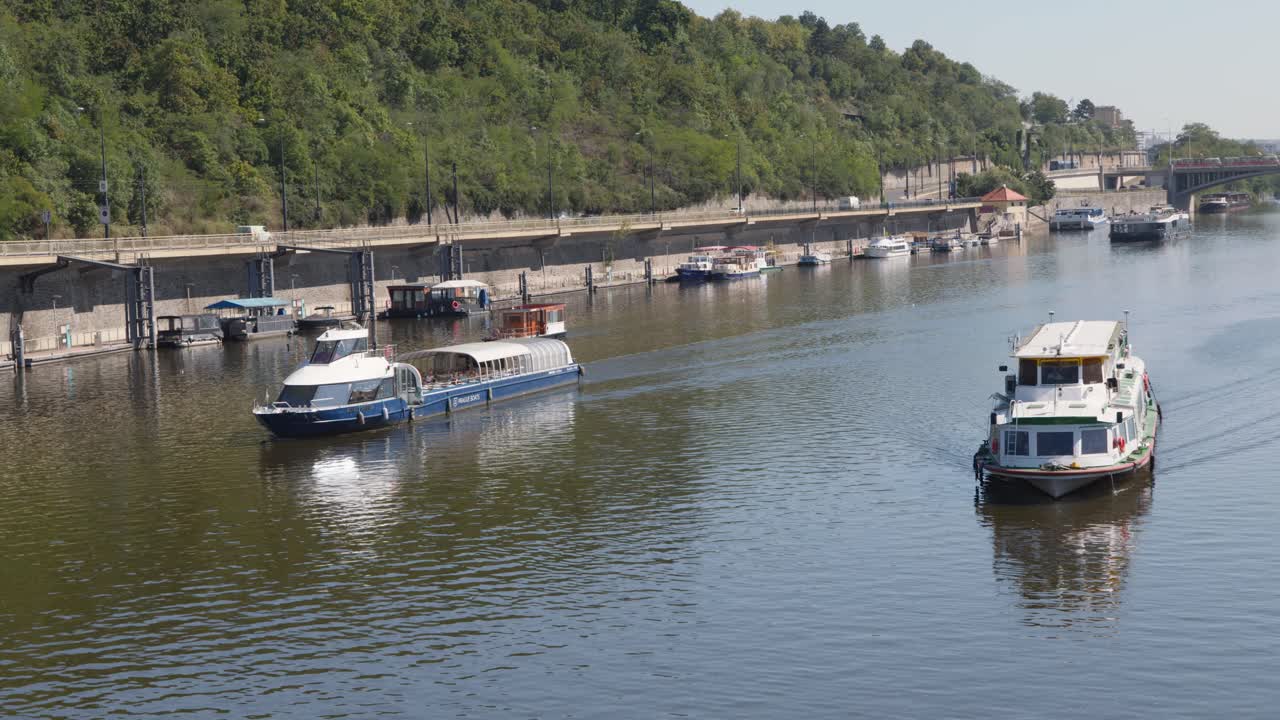 Passenger boats move along Vltava River, sunny day, green riverside, steady wide shot, Prague