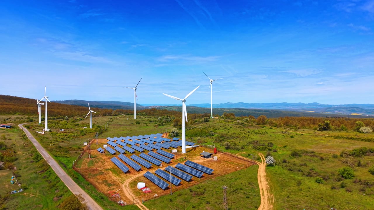 Vista aérea de un parque solar rodeado de aerogeneradores. Caminos de tierra sin pavimentar cruzan el terreno verde y herboso bajo un cielo azul brillante y despejado.