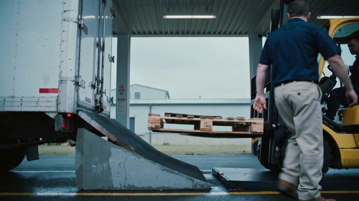 Stepping ramp at covered bay, dock worker in navy polo guiding forklift pallet onto dock plate