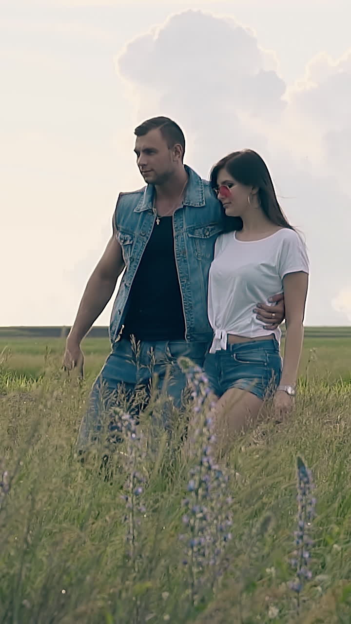 girl in orange sunglasses walks on yellow field with man in denim vest and shorts under white sky