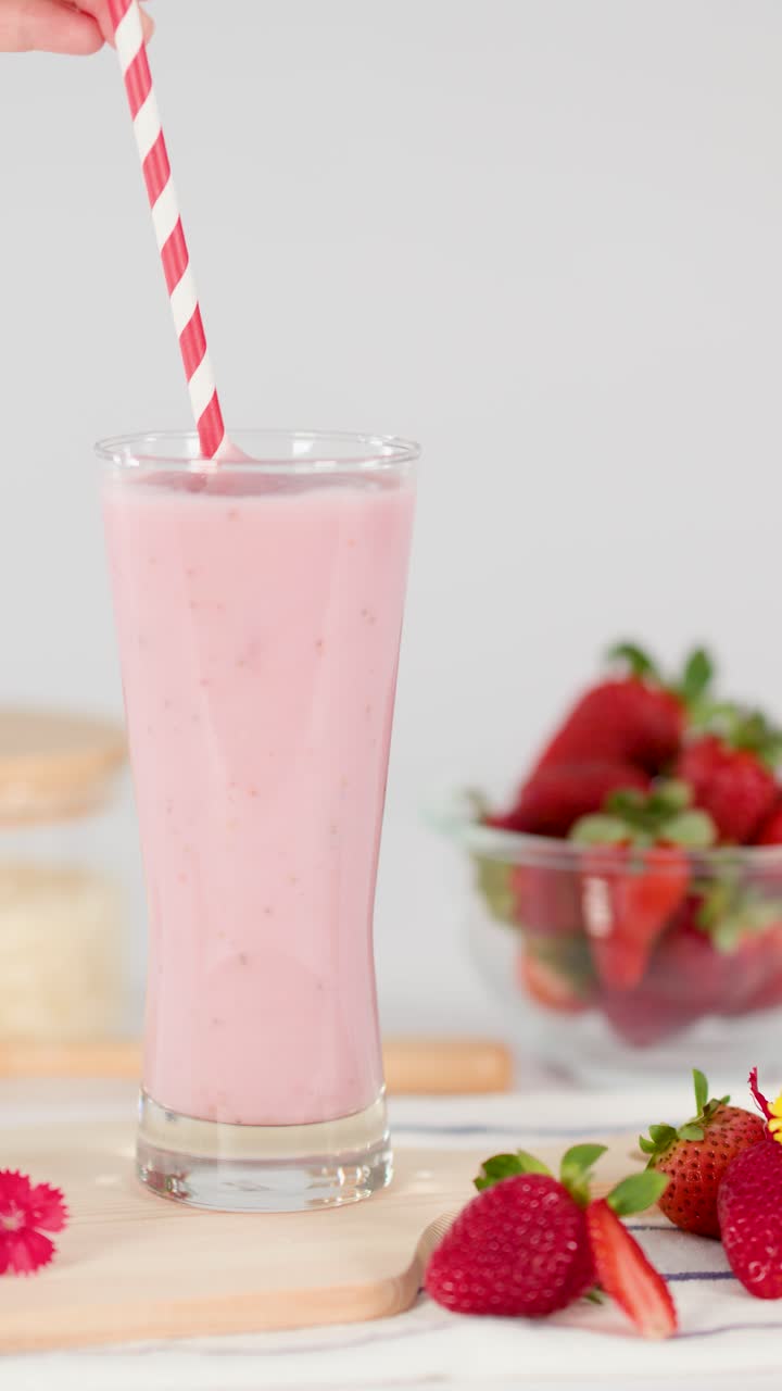 Una mano inserisce una cannuccia a strisce in un cremoso milkshake rosa alla fragola, illuminazione da studio brillante, sfondo di frutta fresca.