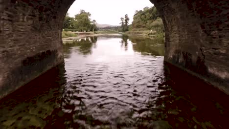 Volando-A-Través-De-Un-Río-Que-Fluye-Y-Volando-Bajo-Un-Viejo-Puente-De-Ladrillo-En-El-Campo-Irlandés