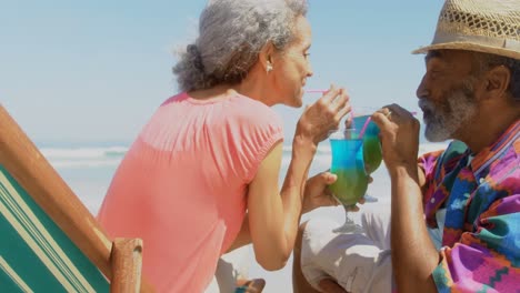 side view of active senior african american couple drinking cocktail on deckchair at beach 4k