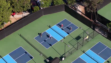 Aerial-view-circling,-looking-down-over-young-adult-foursome-playing-pickleball-on-blue-sunny-tennis-court
