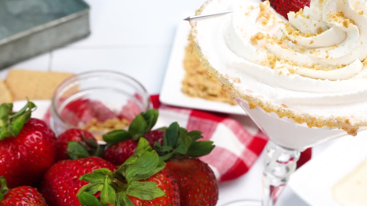 Strawberry Shortcake Martini cocktail with whipped cream, graham cracker crumbs and a fresh red strawberry. Making a cocktail for happy hour. A summer time berry drink with red gingham napkin.
