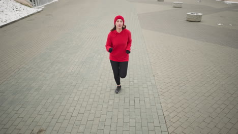 mujer de negocios corriendo en el pavimento entrelazado durante el invierno con cabello fluido y capucha roja vibrante, suelo cubierto de nieve, macetas de flores y elementos urbanos serenos con rieles de hierro