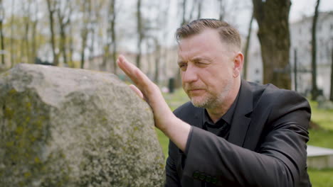 Close-Up-View-Of-Man-In-Black-Suit-Kneeling-In-Front-Of-A-Tombstone-And-Touching-It