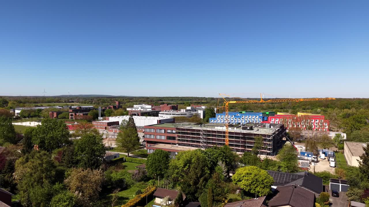 Sorvolo aereo di un cantiere per la costruzione di una scuola di quartiere con gru a torre, circondato da case residenziali, alberi e paesaggio primaverile soleggiato in un'area urbana periferica.