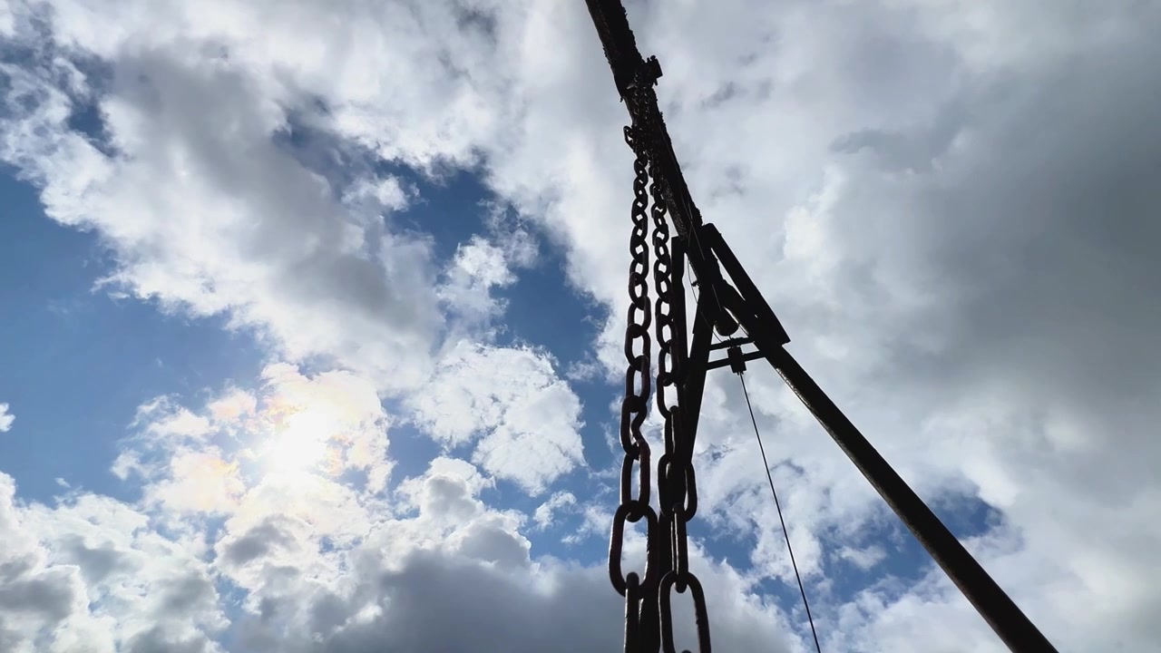 Un treppiede metallico e un meccanismo di verricello a catena visti dal basso contro un cielo nuvoloso e illuminato dal sole. Adatto per attrezzature industriali, costruzioni e concetti meccanici per esterni