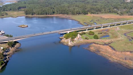 Puente-Con-Coches-Circulando-Sobre-El-Rio-Ulla-Y-Las-Ruinas-De-Las-Torres-De-La-Zona-De-Desembarco-Vikingo-Del-Oeste,-Tarde-Soleada,-Disparos-De-Drones-Avanzando-Cenital-2