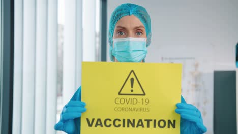 Close-Up-Of-Young-Pretty-Female-Healthcare-Worker-Medic-In-Protective-Suit-And-Medical-Mask-Holding-In-Hands-Yellow-Paper-Card-With-Notification-About-Covid-19-Coronavirus-Vaccination