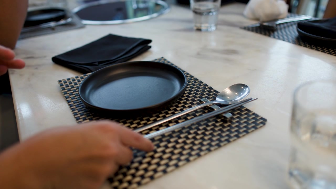 Close-up shot of hands placing metal utensils on a woven placemat in a restaurant