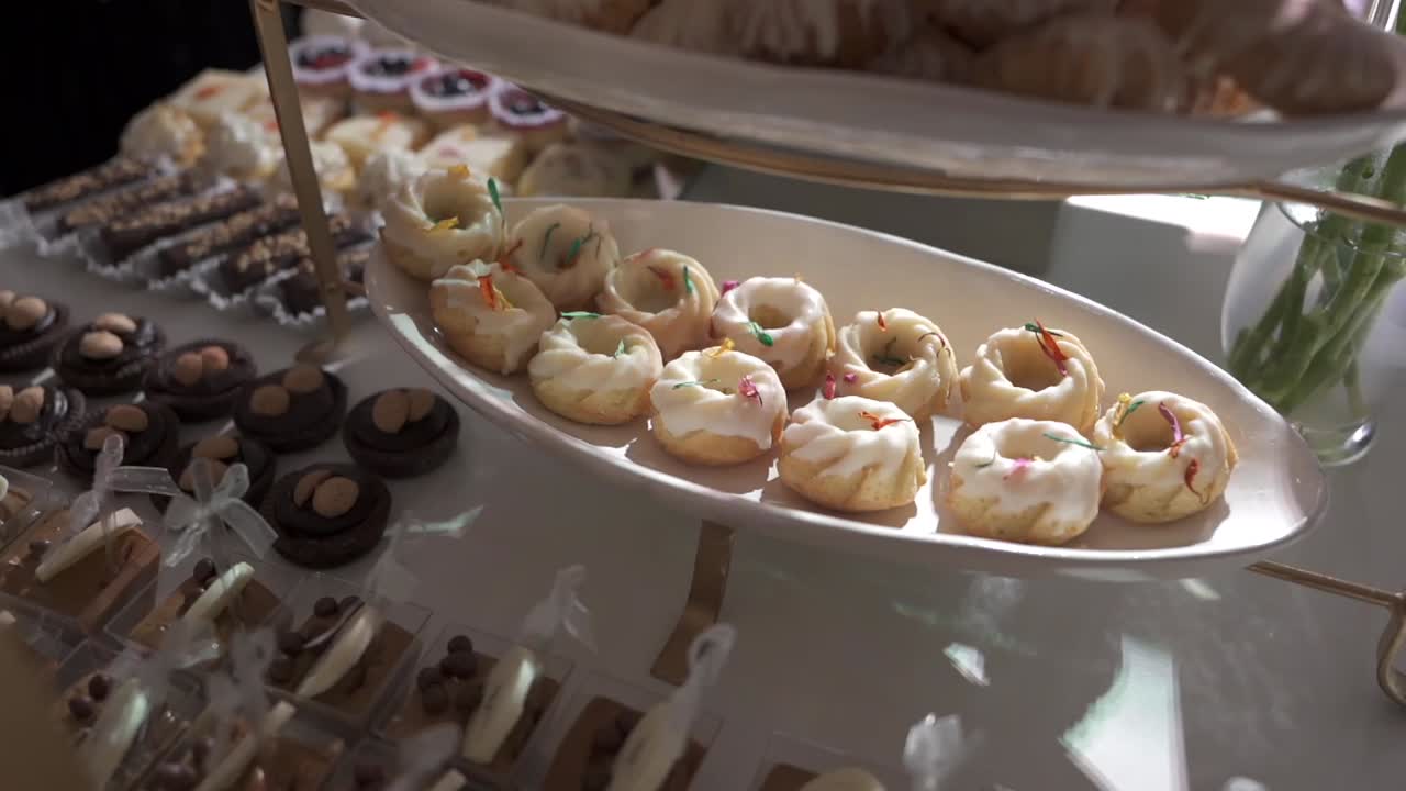 Slow motion optagelse i kredsløb omkring et elegant dessertbord fyldt med et udvalg af mini-desserter og kager arrangeret på etagestande i blødt lys, hvilket skaber en luksuriøs buffet.