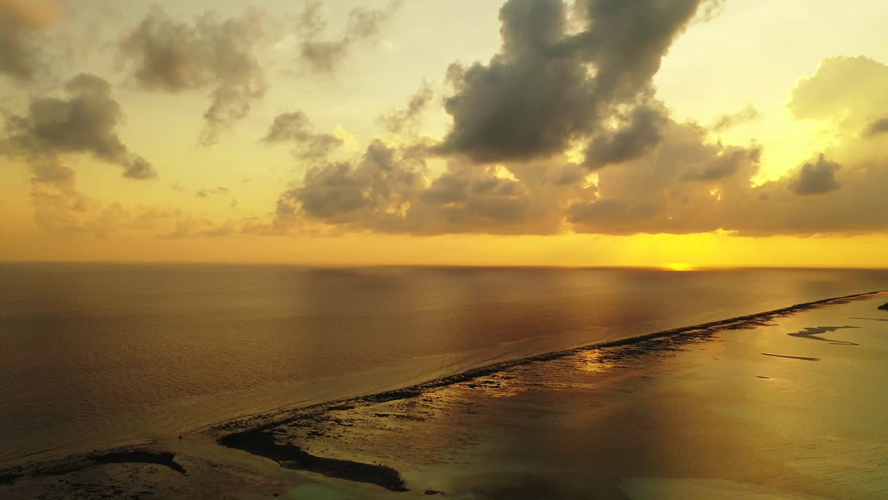 Beautiful sunset at golden hour with yellow sky and grey clouds reflecting over endless ocean and sandy stripe beach in Maldives