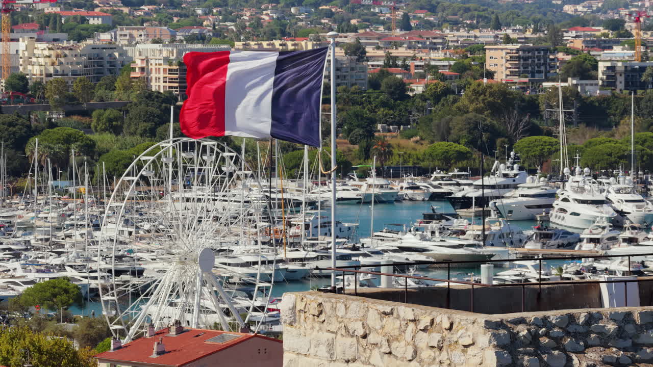 Aerial drone view of the French flag waving above Antibes marina, with luxury yachts, sailboats, and a Ferris wheel in the background