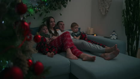 caucasian family sitting together on a sofa and watching tv at home during christmas holidays decorated apartment interior. high quality 4k footage