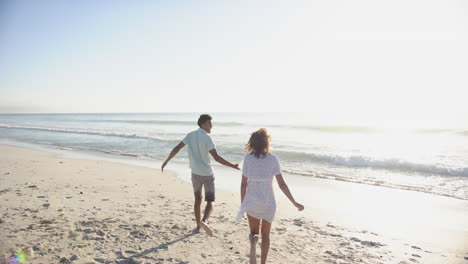 Ein-Gemischtrassiges-Paar-Läuft-An-Einem-Sonnigen-Strand-Entlang,-Mit-Kopierraum,-Den-Rücken-Zur-Kamera