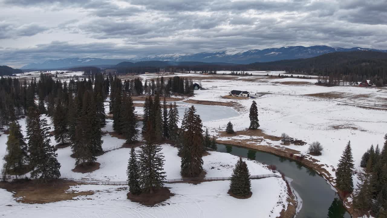 Riprese aeree con drone di una penisola e un lago innevati nel Montana, con gruppi di alberi sempreverdi circondati da acqua parzialmente ghiacciata e montagne distanti sotto un cielo nuvoloso