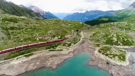 vista aérea con un avión no tripulado de un tren rojo que pasa cerca de un lago azul en la base de una montaña en suiza. hermoso paisaje bajo el cielo azul 4k