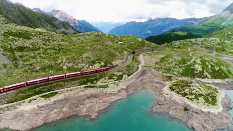 aerial view with a drone of a red train passing near a blue lakeat the bottom of a mountain in switzerland . beautiful landscape under the blue sky 4k