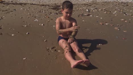 caucasian toddler boy in a swimsuit, playing with sand, in sandy beach of varkiza, attica, greece