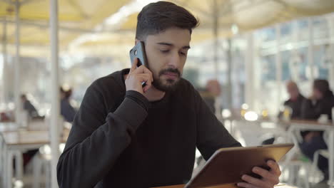 Serious-young-man-talking-by-smartphone-and-using-tablet-pc