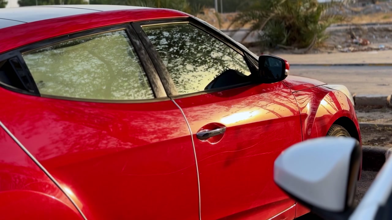 Un'auto rossa parcheggiata su una strada esterna soleggiata. La calda luce solare diurna riflette gli alberi nei finestrini laterali oscurati, con uno specchietto retrovisore bianco sfocato in primo piano