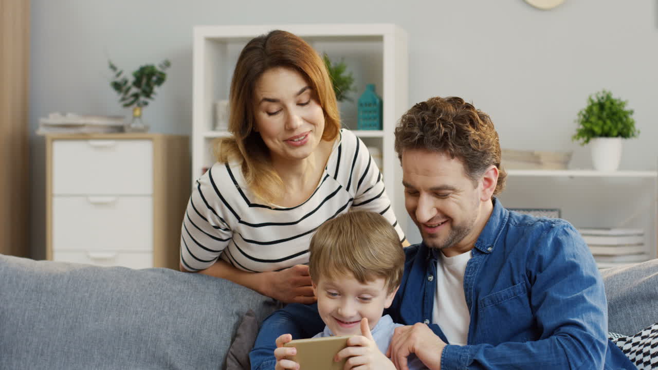 niño lindo sentado en el sofá de la sala de estar y jugando juegos en el smarthone, sus padres mirándolo