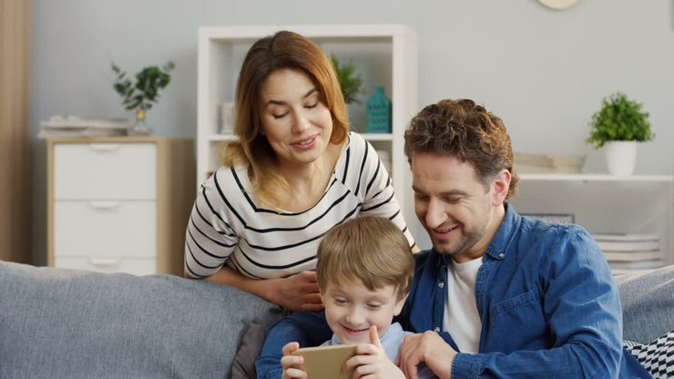 niño lindo sentado en el sofá de la sala de estar y jugando juegos en el smarthone, sus padres mirándolo