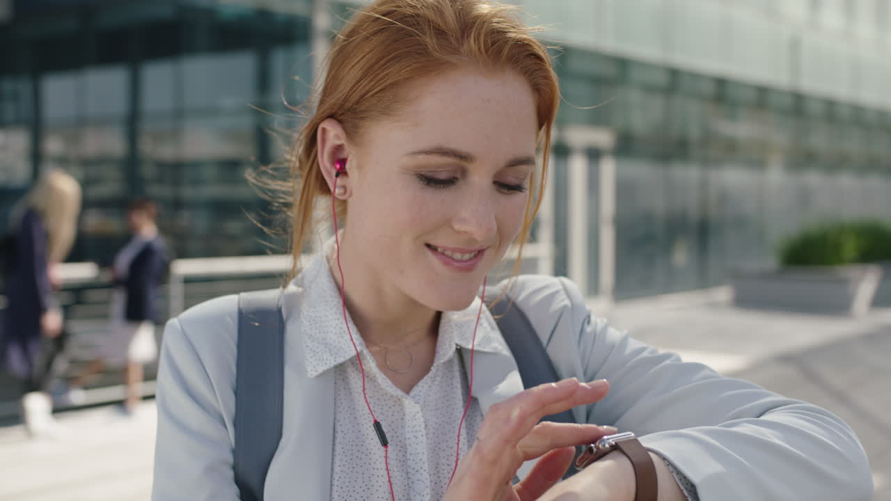 retrato en primer plano de una feliz mujer de negocios de cabeza roja que lleva auriculares escuchando música navegando mensajes usando un reloj inteligente