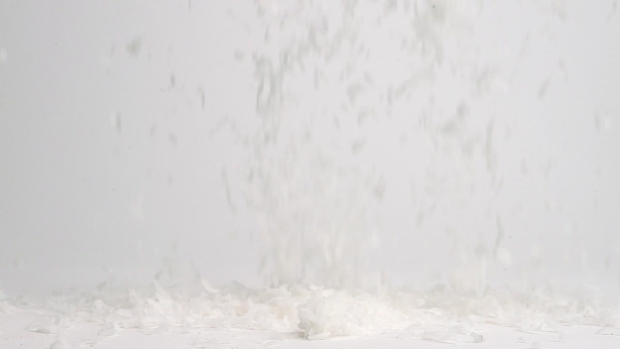 흰색 코코넛 플레이크가 흰색 테이블 위에 비가 내리고 슬로우 모션으로 더미에 착륙합니다.