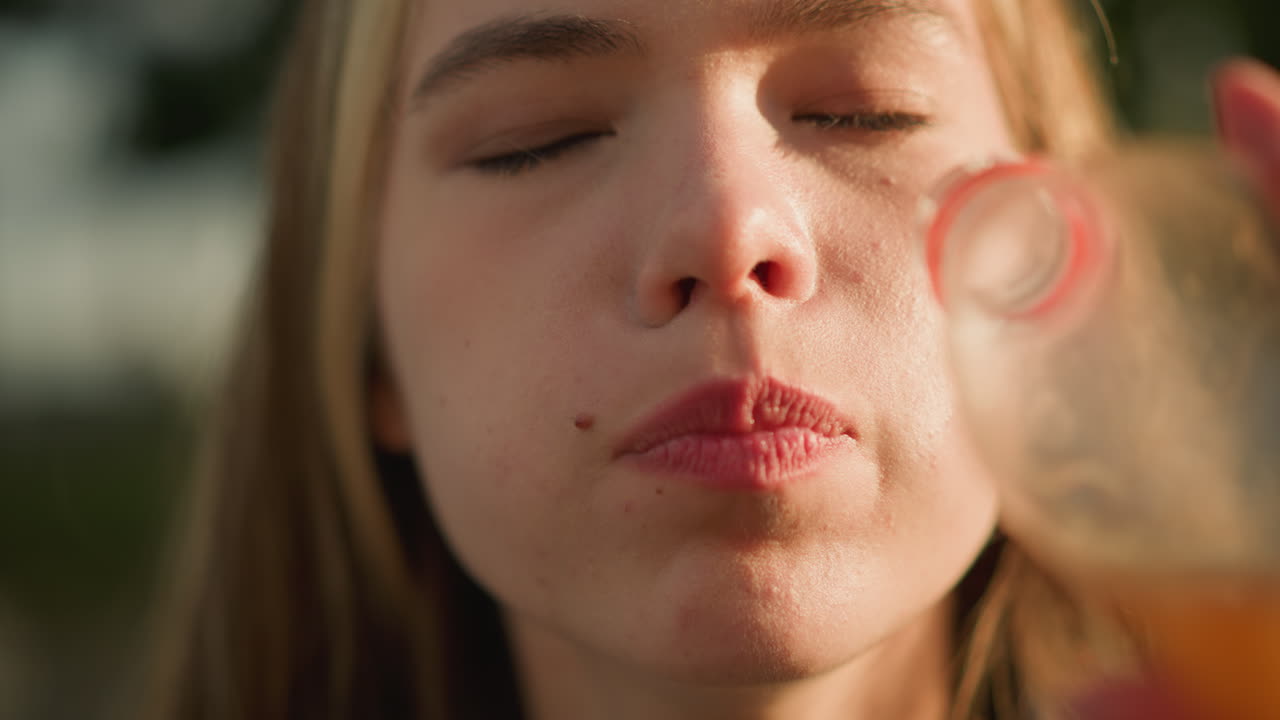 야외에서 플라스틱 병에서 오렌지 주스를 마시는 여성의 클로즈업, 그녀의 얼굴과 병에 빛이 빛나고, 빛과 초록색의 부드러운 배경 빛, 만족을 반영합니다.