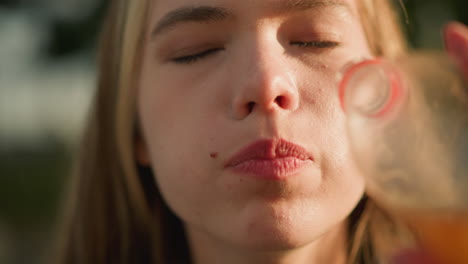 close-up of woman drinking orange juice from plastic bottle outdoors, with sunlight shining on her face, bottle, and soft background glow of light and greenery, reflecting satisfaction