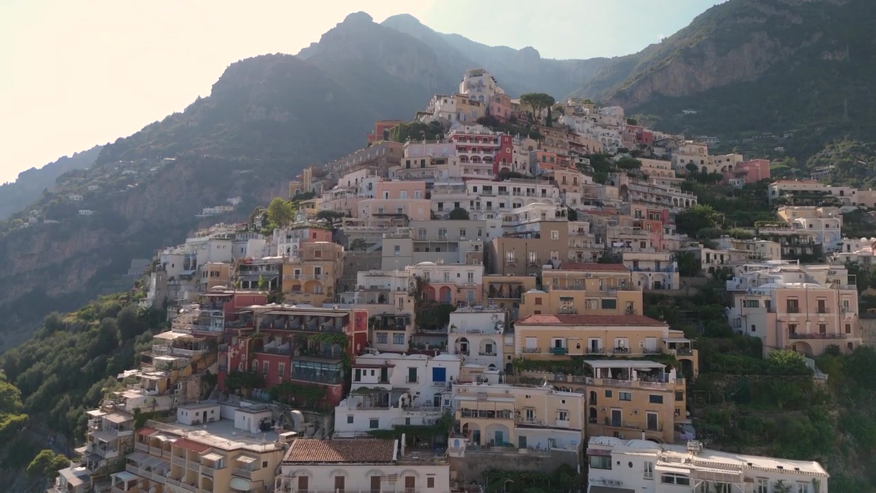 Questa è una vista di Positano, una città sulla Costiera Amalfitana in Italia. La telecamera riprende case colorate disposte a cascata su una collina. Le montagne circondano la città e il cielo è limpido.