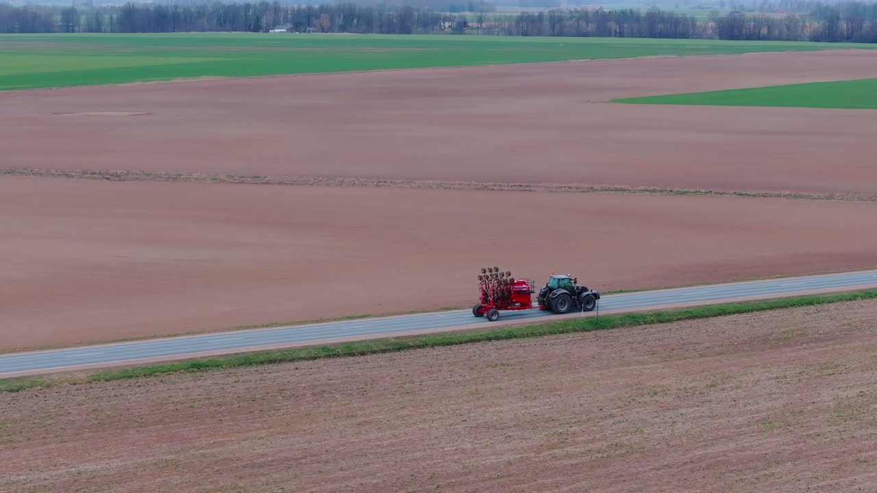 Overcast Baltic landscape as tractor hauls red seeding equipment, empty fields
