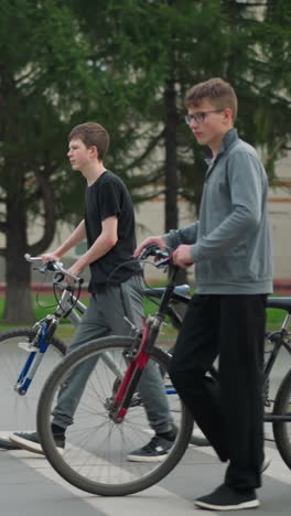 two friends walking side by side with their bicycles across a pedestrian crossing, a blurred view of three people walking in the background, with trees and residential buildings visible