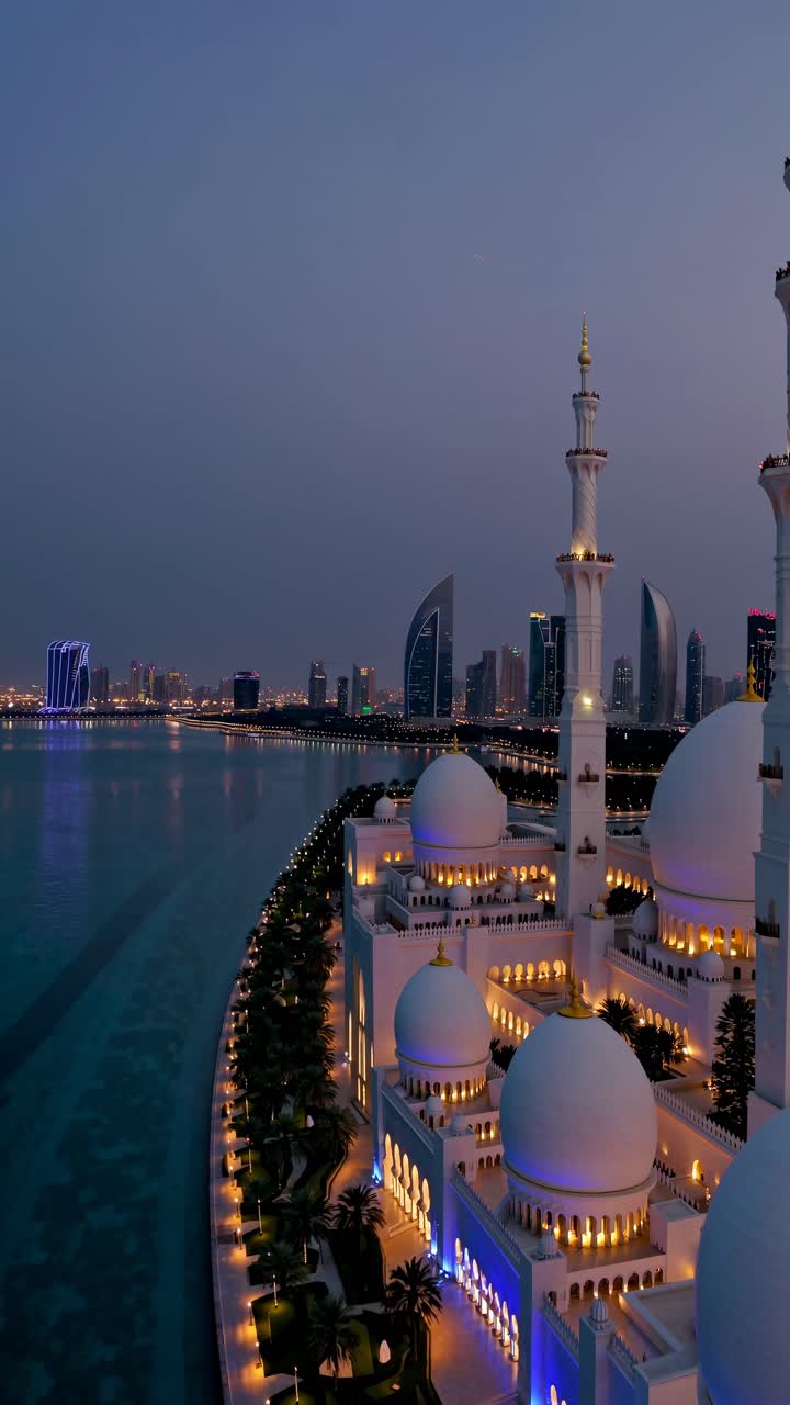 Veduta aerea di una moschea illuminata al crepuscolo con uno skyline cittadino sullo sfondo.