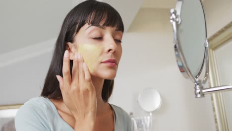 caucasian woman looking in mirror and using cream on face in bathroom