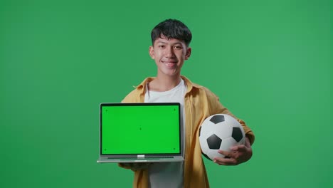 niño asiático con una pelota que muestra una maqueta de portátil de pantalla verde y sacudiendo la cabeza mientras anima al fútbol en el fondo de la pantalla verde