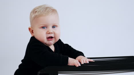 beautiful blond baby boy in black sweater stands by the metal cage. lovely grey-eyed child at white backdrop.