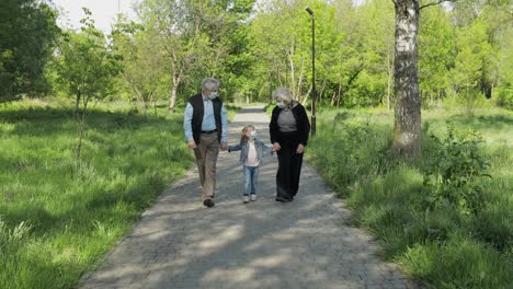 Old-grandparents-with-granddaughter-in-medical-masks-walk-in-park.-Coronavirus