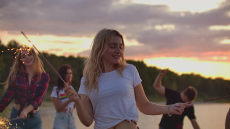 the blonde girl with nude waist is dancing with big bengal lights in her hands on the sand beach with her friends. this is wonderful summer evening on the open air party.