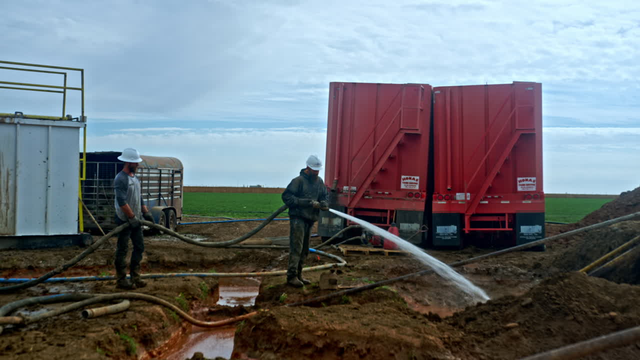 Two workers wearing protective casques hold the pipe hose with water pouring from. Location for production of oil and gas.