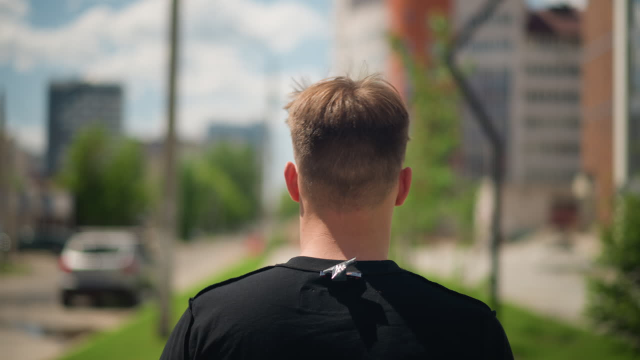 Hombre blanco caminando junto a edificios modernos de cristal, vista trasera que destaca el corte de pelo y los hombros relajados, horizonte de la ciudad y ventanas reflectantes que forman un nítido telón de fondo urbano para un paseo tranquilo