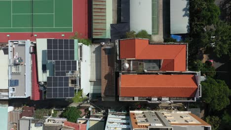Aerial-top-down-tracking-shot-over-modern-rooftops-with-solar-panels,-red-and-green-tennis-courts-and-small-canal-with-traditional-waterfront-houses