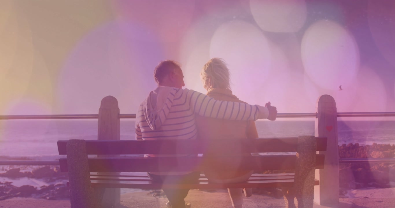 Travel couple sitting bench facing sea man turning lifting phone reaching woman loving under bokeh