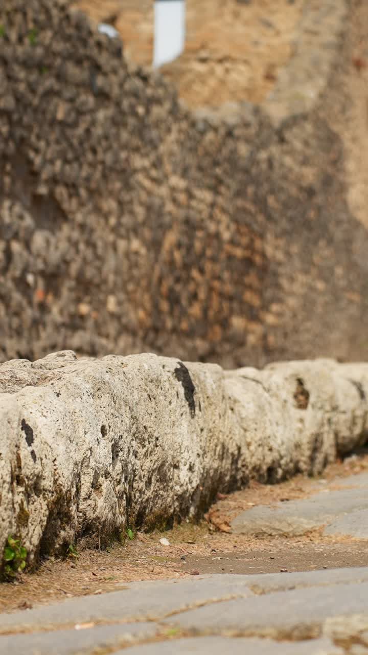 Primo piano verticale di un cordolo in pietra consumato di Pompei e del bordo di un'antica strada, che evidenzia i dettagli della costruzione stradale romana e le texture consumate nel sito archeologico.