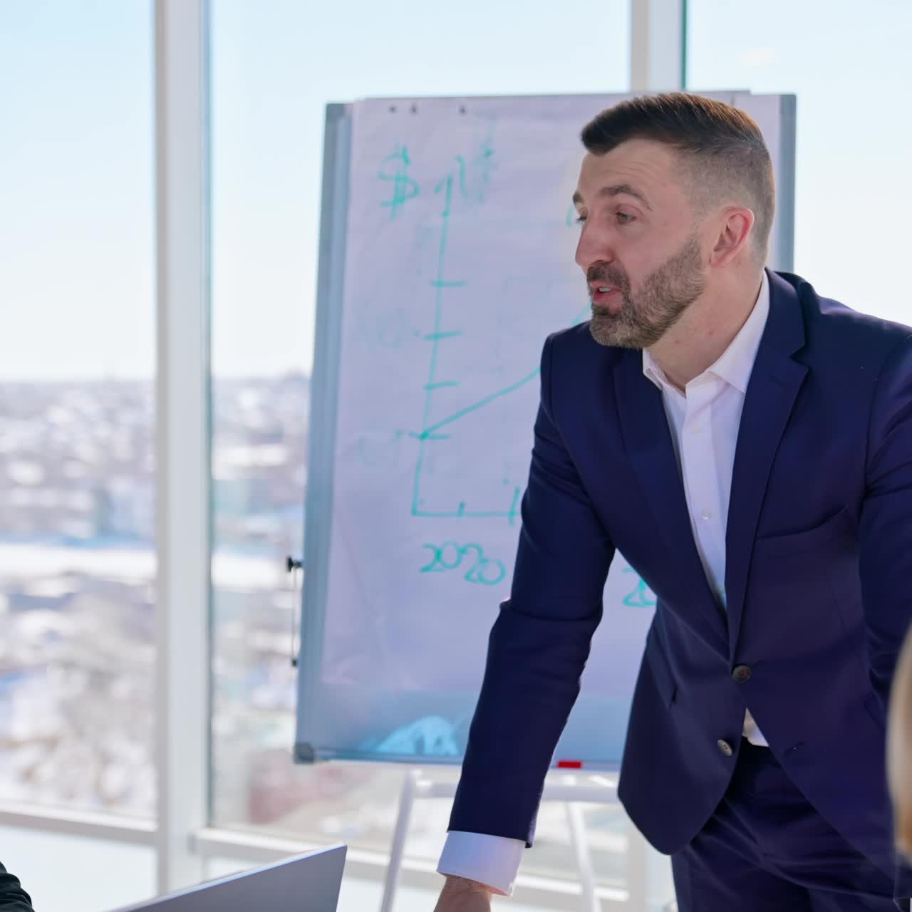 Business conversation in office. Business colleagues sit at table and listen to entrepreneur in elegant suit on the large window background with a city view