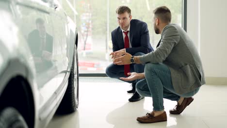 two men cheerful customer and friendly salesman are talking and gesturing discussing car model while squatting near expensive automobile. buying and selling vehicles concept.