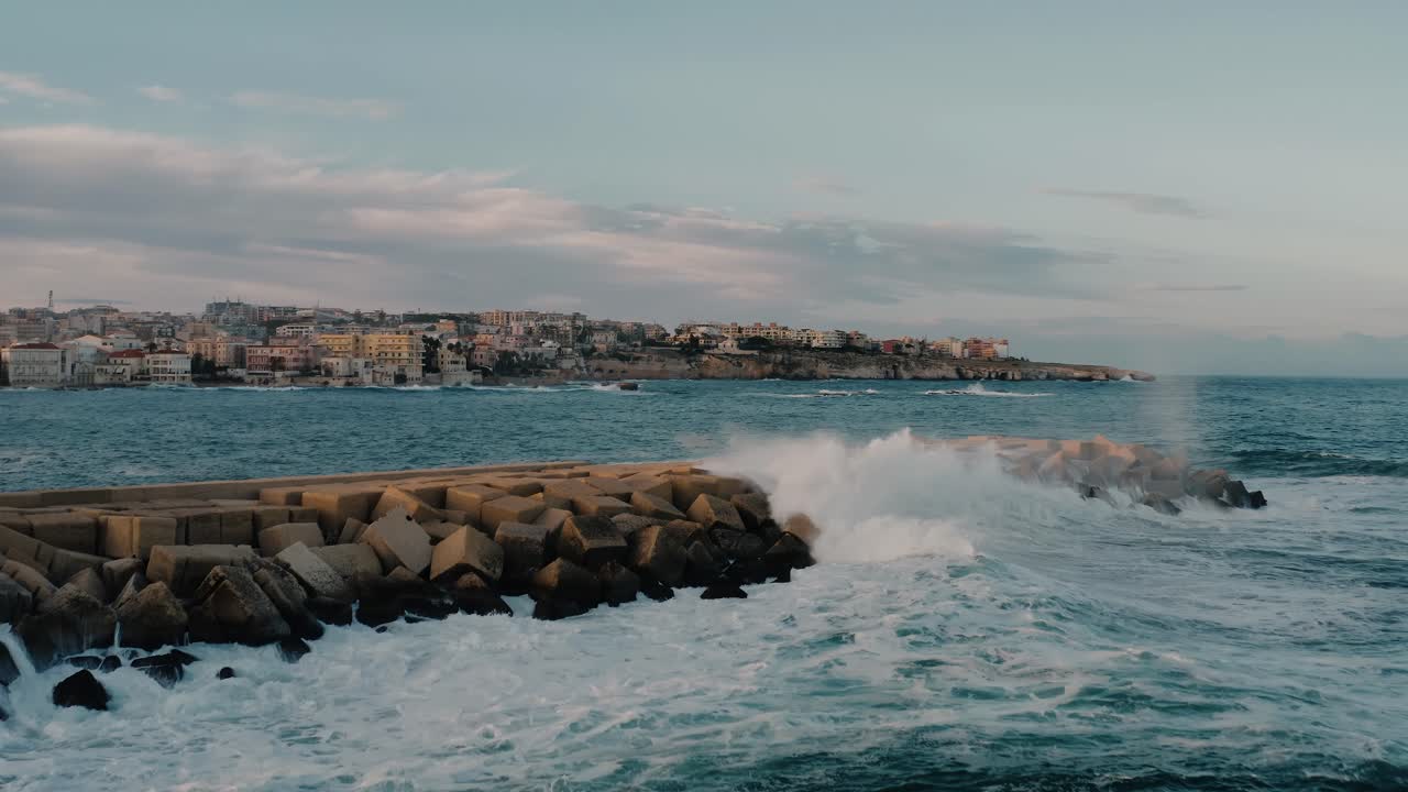 una gran ola chocando contra el muelle construido de piedras con una ciudad de siracusa en el fondo. paisaje de sicilia.